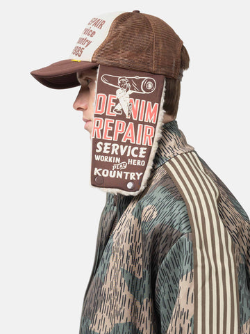 Side view of model wearing the KAPITAL DENIM REPAIR SERVICE LUMBER Trucker Cap ECTxBrwn with ear flap down